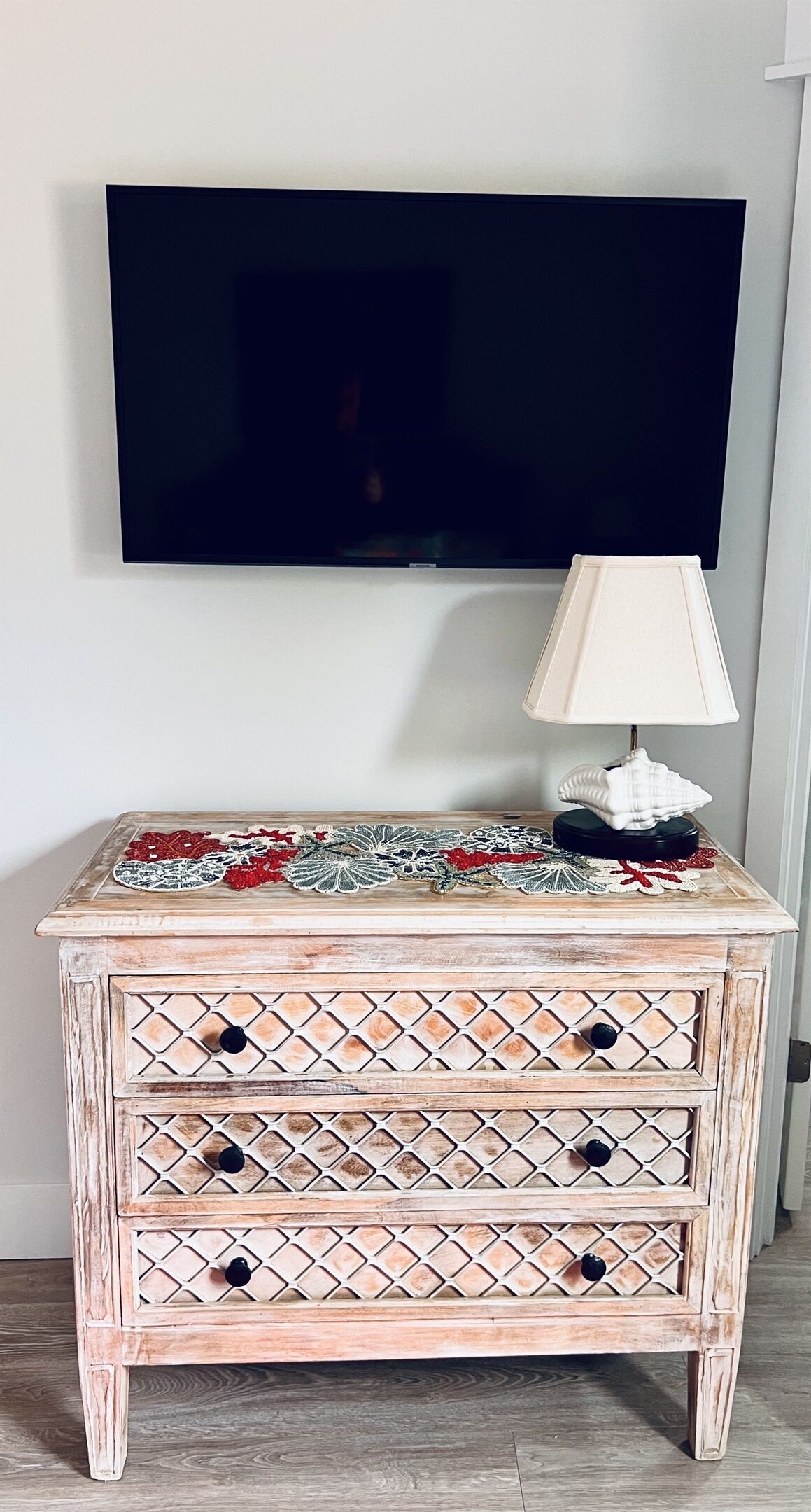 Une commode en bois, ornée d’un plateau décoratif, avec une lampe et un téléviseur accroché au mur derrière.
