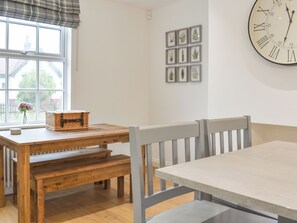 Un coin repas avec une table et un banc en bois, une horloge au mur et des reproductions botaniques encadrées.