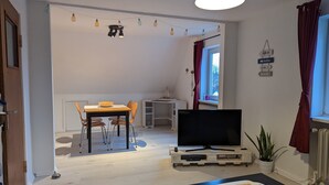 A living room with a TV on a wooden pallet, a dining table with chairs, a potted plant, and a string of lights. - Vacation apartment near the beach with sea view (Hohwacht (Ostsee))