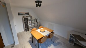 A dining area with a table and chairs, a ceiling light fixture, a wooden shelf unit with decorative items, and a door in the background. - Vacation apartment near the beach with sea view (Hohwacht (Ostsee))