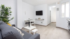 A modern living room with a grey sofa, a small table, a potted plant, a flat-screen TV mounted on the wall, and a kitchen area with a white countertop and cabinets. - Seafront San Andres (Santa Cruz de Tenerife)