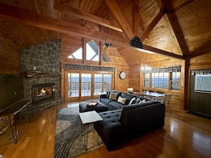 A cozy log cabin interior with a stone fireplace, a black leather sofa, wooden floors, and large windows offering mountain views.
