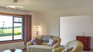 Un salon avec un canapé, un fauteuil et une petite table sur laquelle se trouve un bol. On y trouve également une lampe, une commode et une fenêtre avec des rideaux.