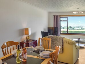 Un coin repas avec une table dressée pour deux personnes, un canapé, un téléviseur et une fenêtre donnant sur la campagne.