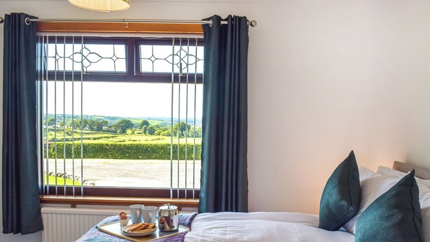 Une chambre avec un lit, un plateau contenant des aliments pour le petit-déjeuner et une vue sur un paysage verdoyant par une fenêtre ornée de rideaux.