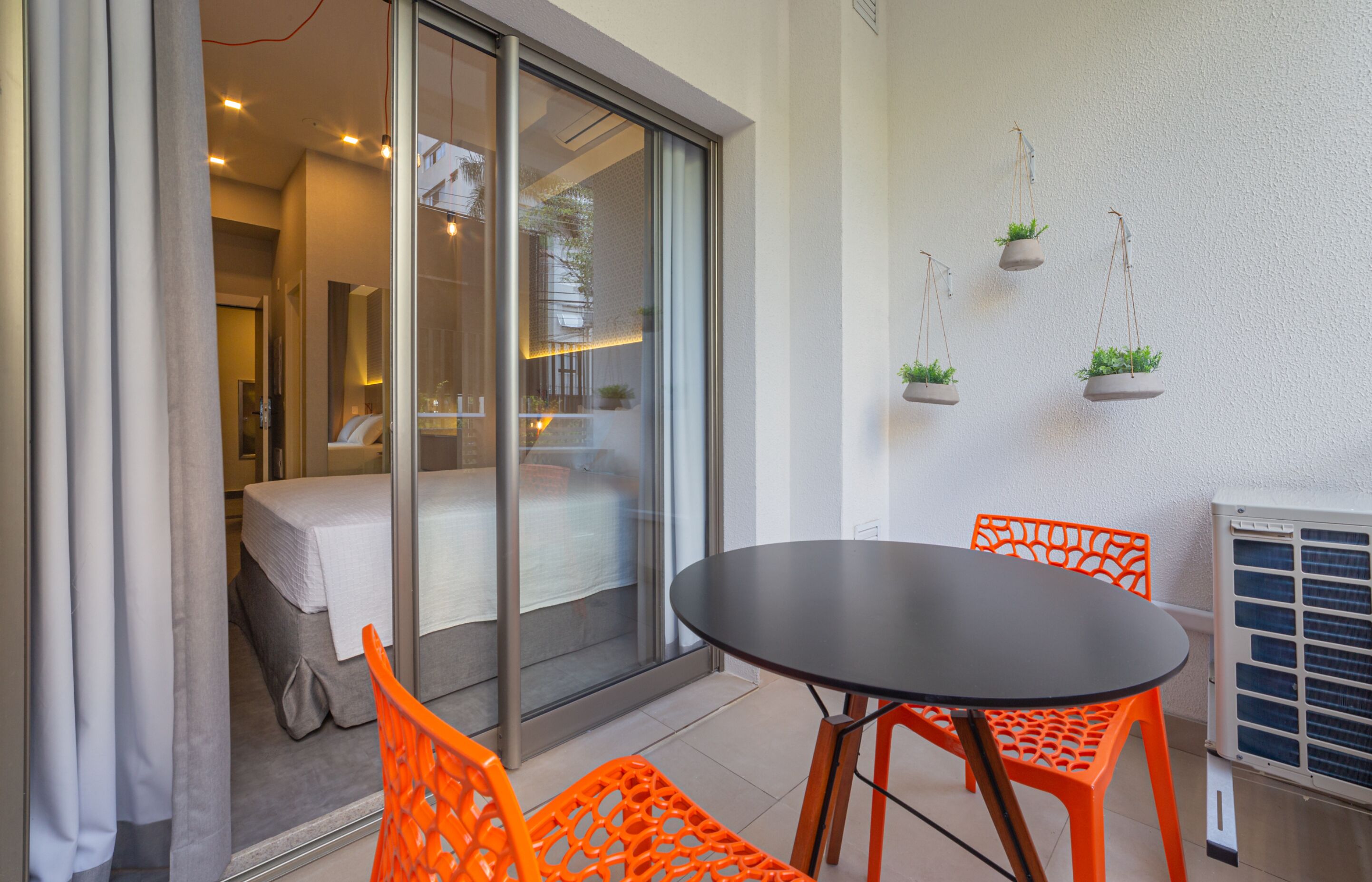 A modern hotel room with a sliding glass door leading to a balcony, a small round table, and orange chairs.