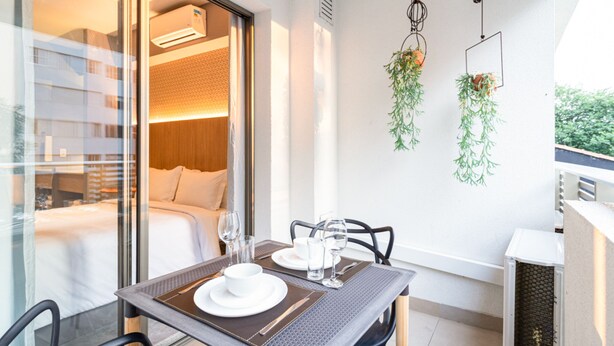 A modern hotel room with a glass door leading to a balcony, a dining area with a table set for two, and a bed with a wooden headboard. - Charlie Girassol Vila Madalena (São Paulo)