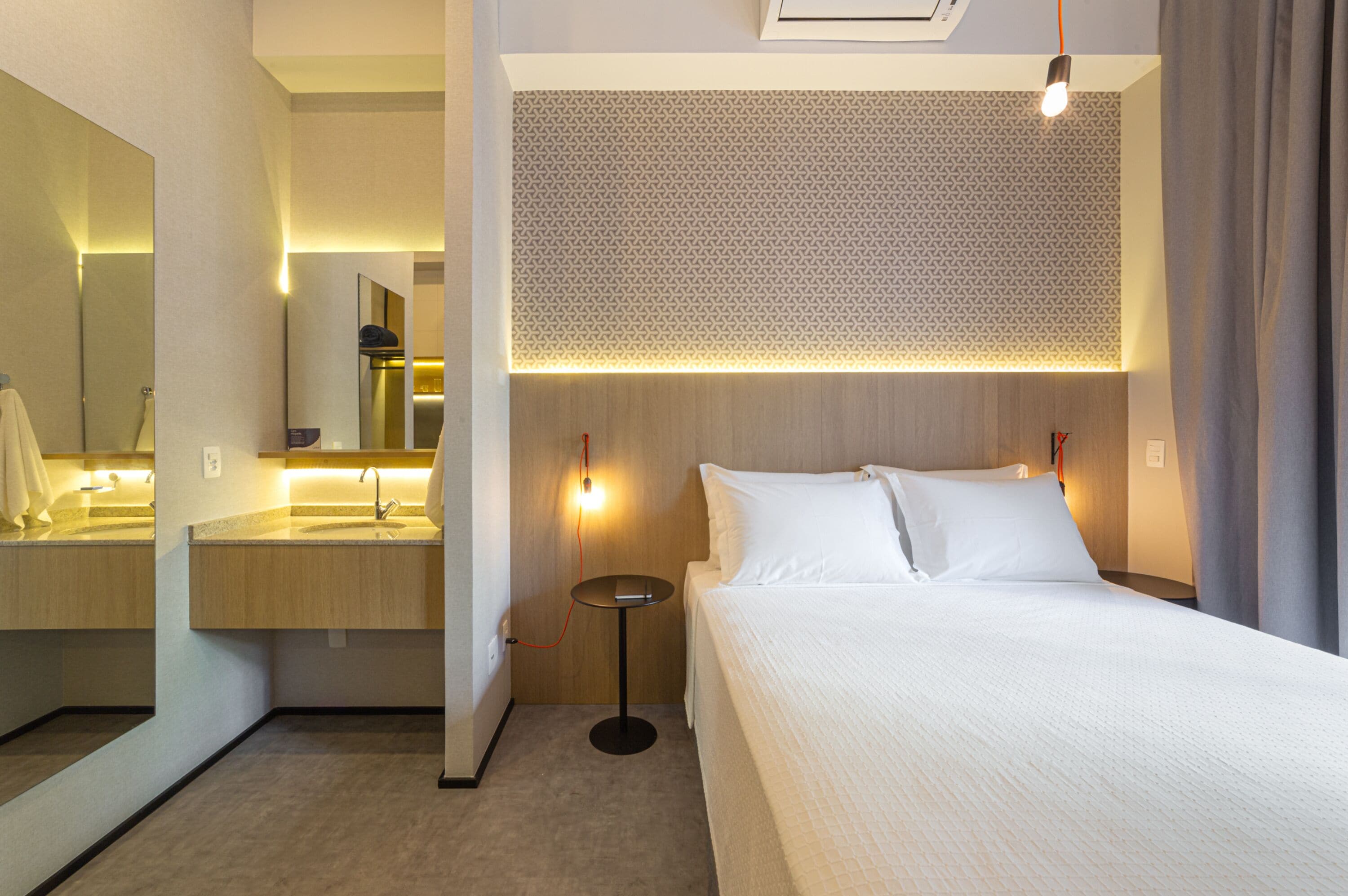 A hotel room with a bed, a bedside table, a mirror, a sink, and a wall-mounted light fixture.