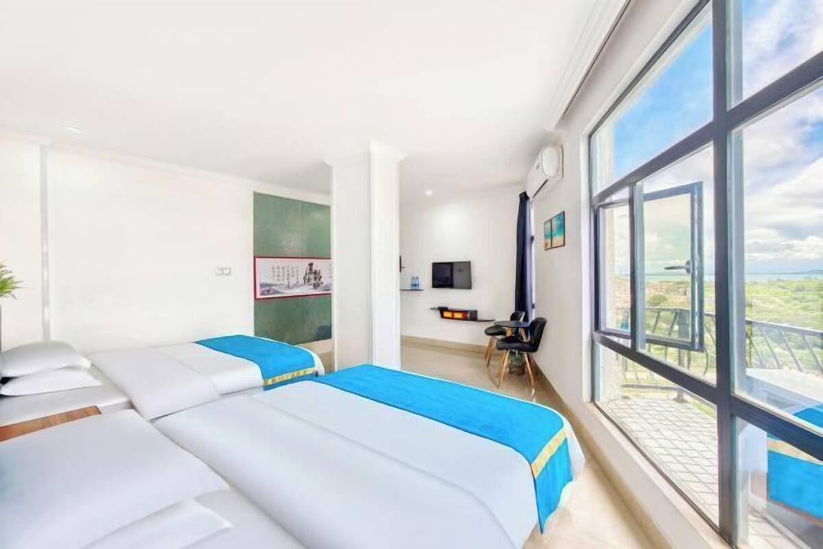 A hotel room with two beds, a desk, a chair, a TV, and large windows.