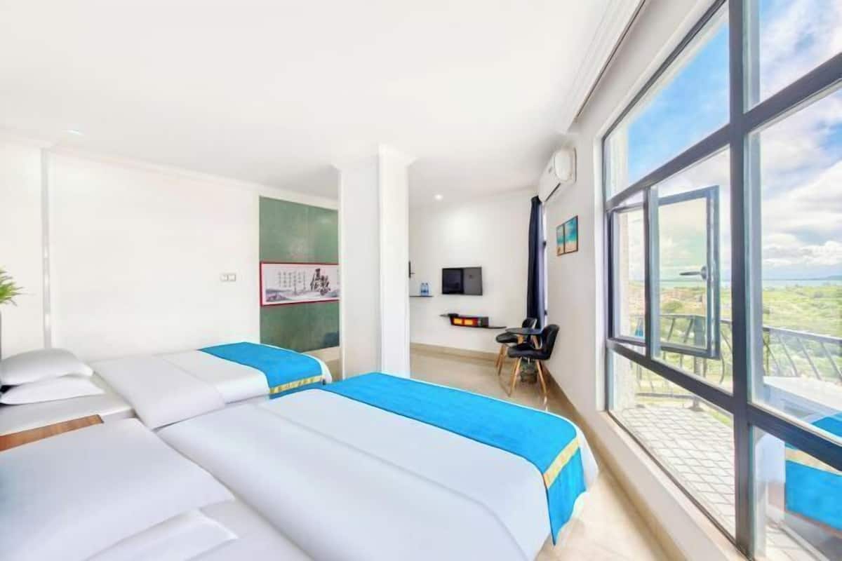 A hotel room with two beds, a desk, a chair, a TV, and large windows.