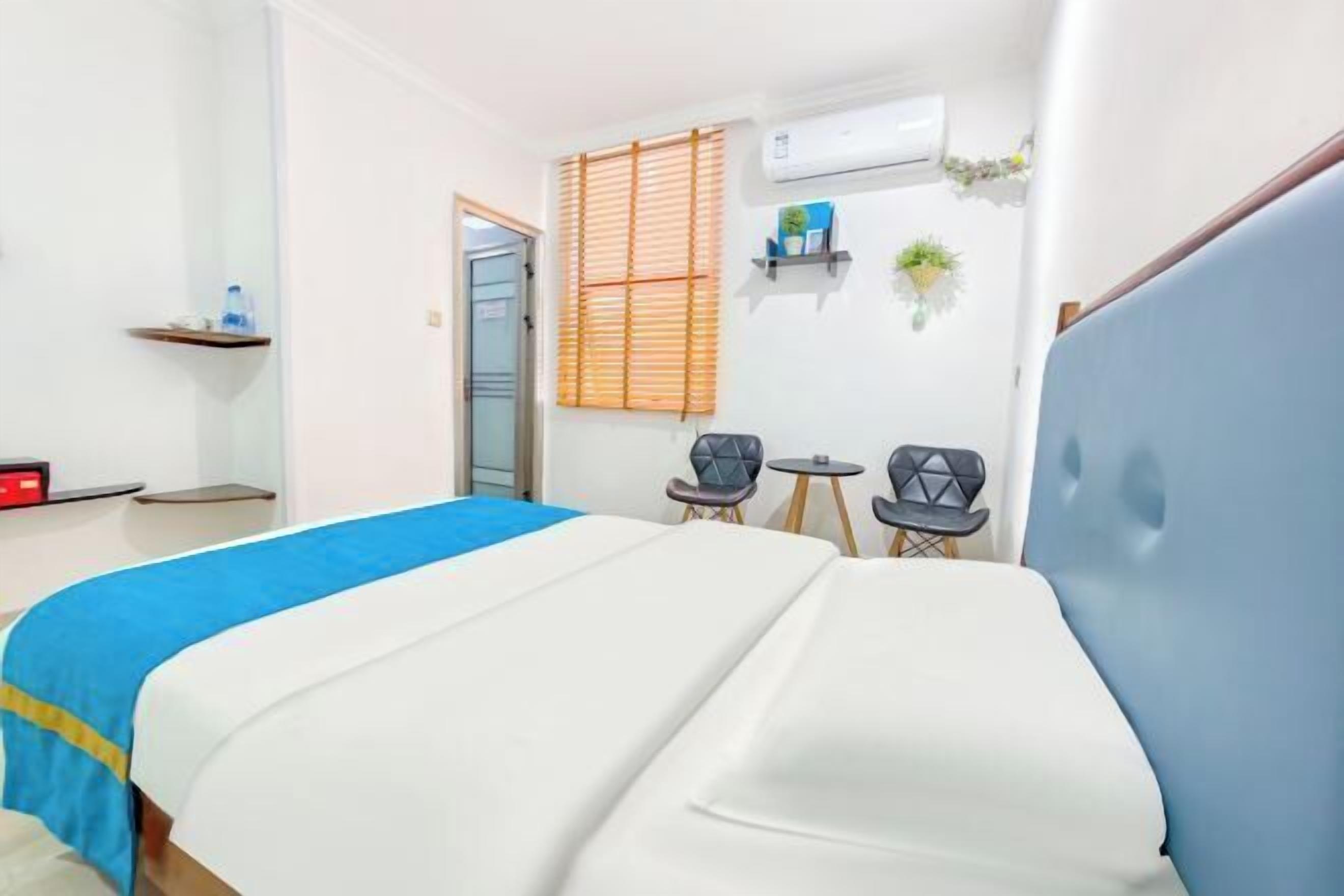 A hotel room with a bed, two chairs, a small table, a shelf with items, and an air conditioning unit.