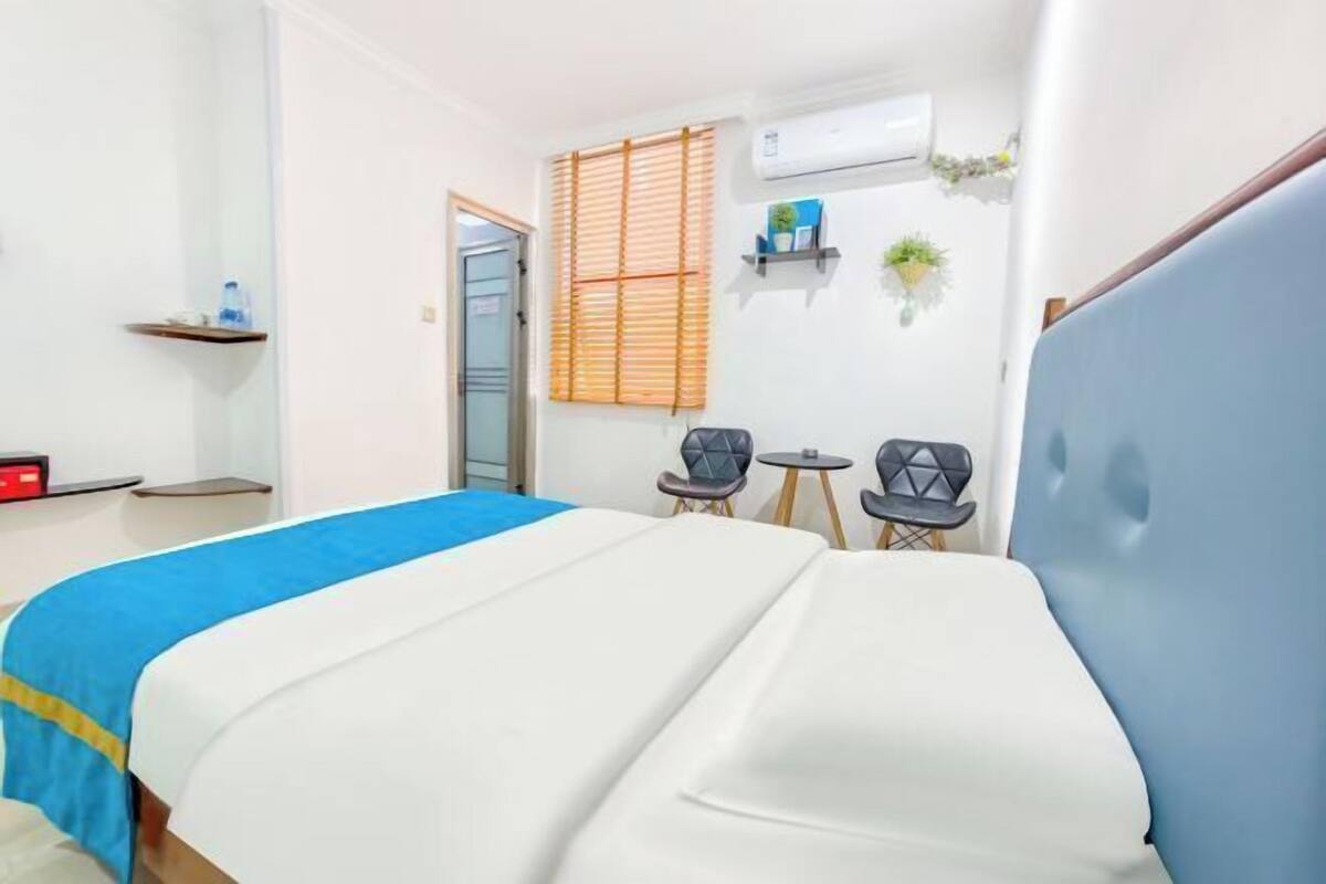 A hotel room with a bed, two chairs, a small table, a shelf with items, and an air conditioning unit.