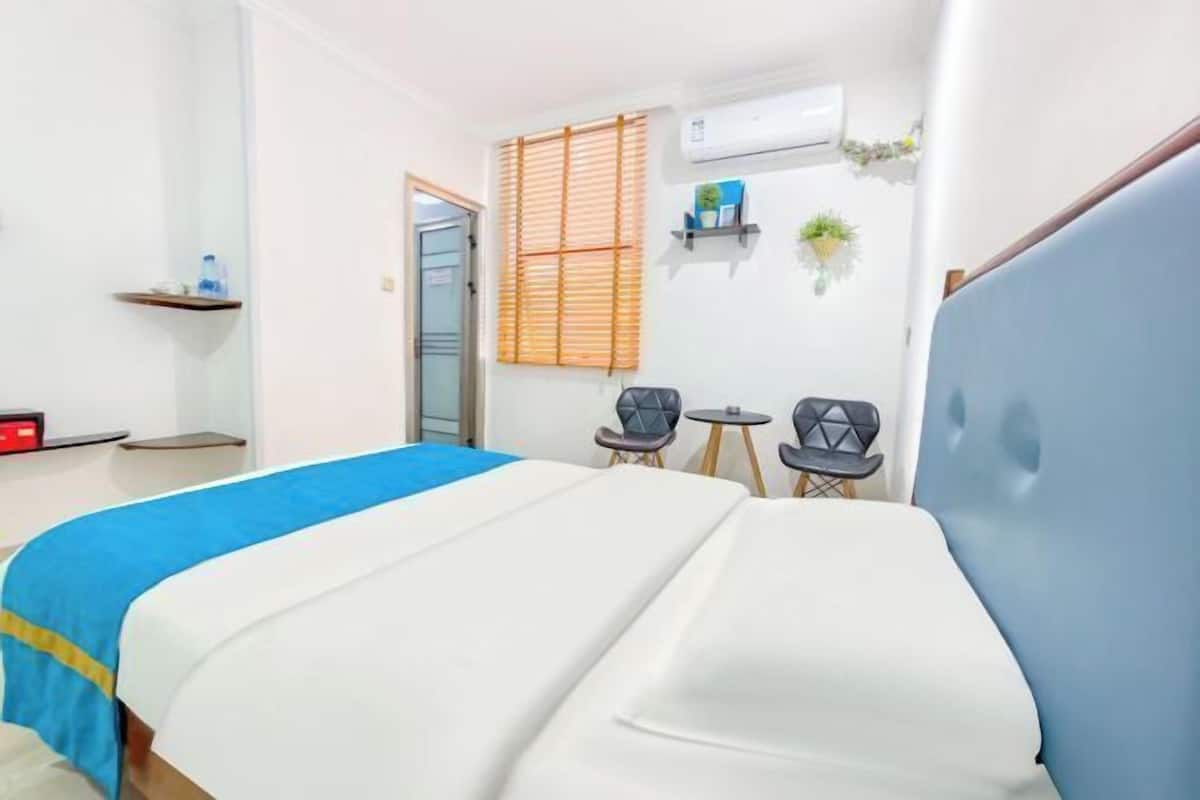 A hotel room with a bed, two chairs, a small table, a shelf with items, and an air conditioning unit.