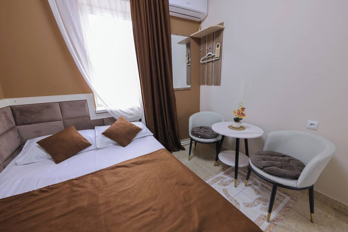 Un cuarto de hotel con una cama, dos sillas, una mesita con un florero y un espejo en la pared.