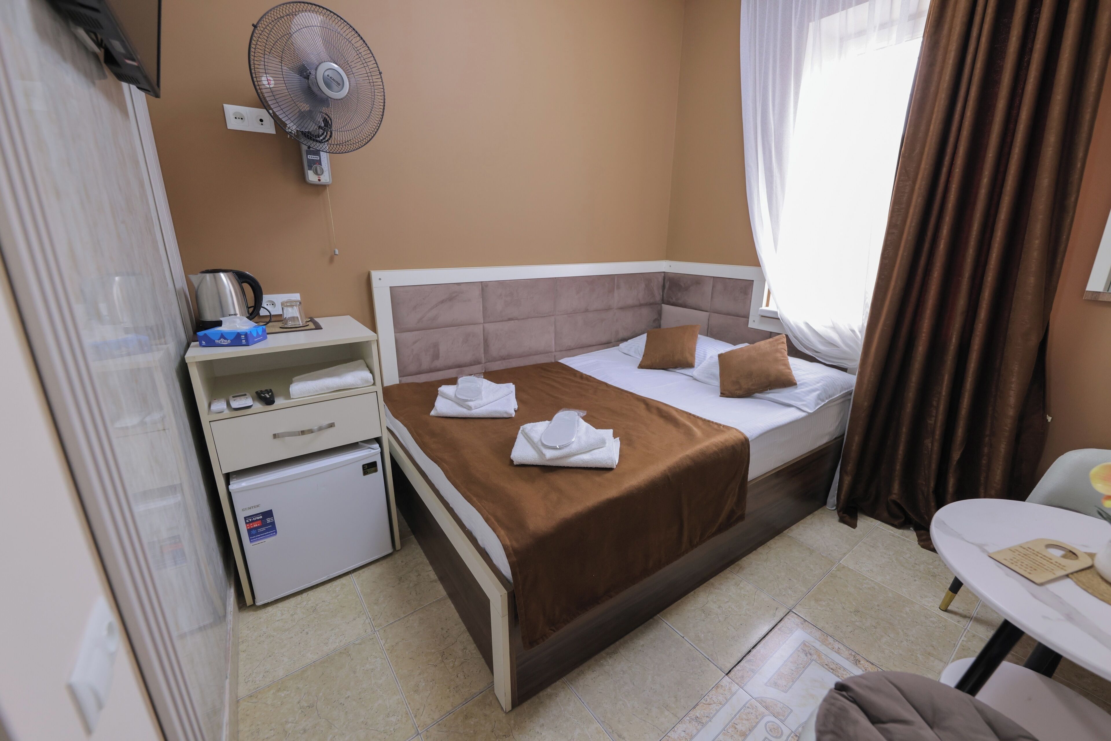 A hotel room with a bed, a bedside table, a small fridge, a fan, and a window with curtains.