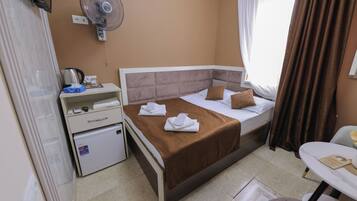 A hotel room with a bed, a bedside table, a small fridge, a fan, and a window with curtains.