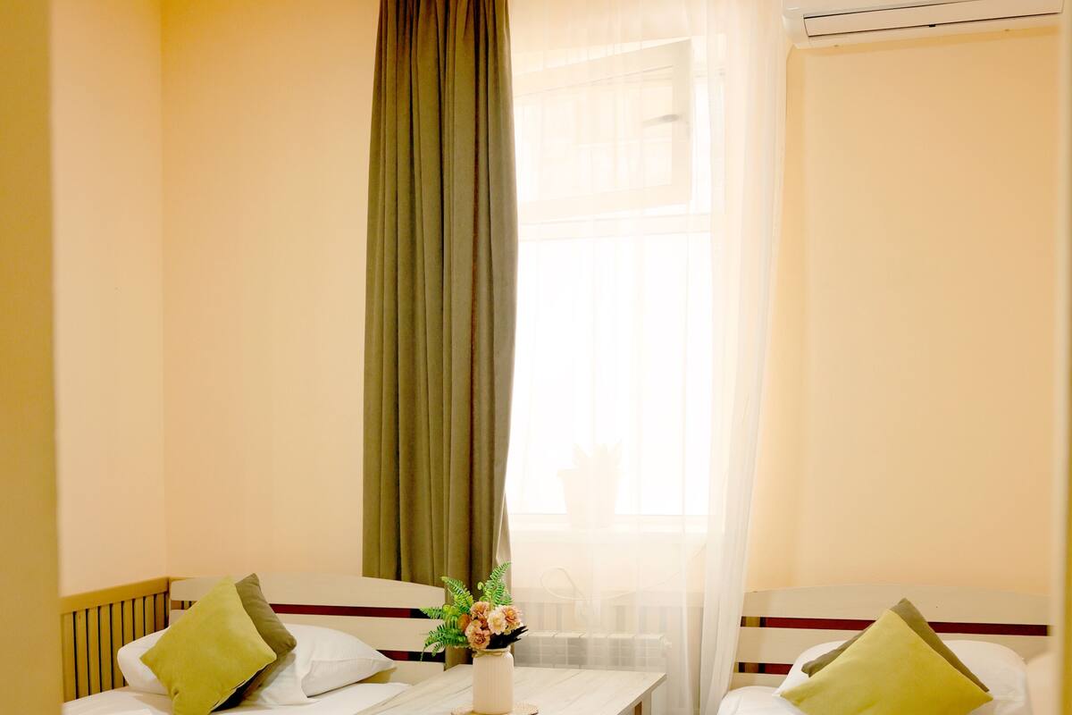 Habitación de hotel con dos camas, una mesa, un ventanal con cortinas y un equipo de aire acondicionado.