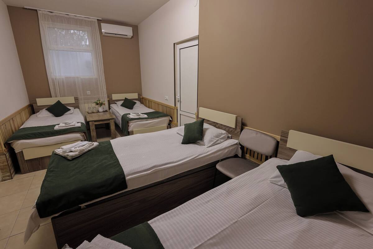Habitación de hotel con cuatro camas individuales, una mesita con lámpara, una silla y una ventana con cortinas traslúcidas.