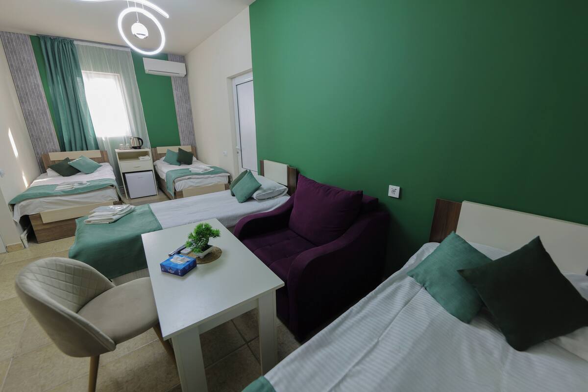 Habitación de hotel con dos camas, una mesita con una planta, un sofá morado y una silla.