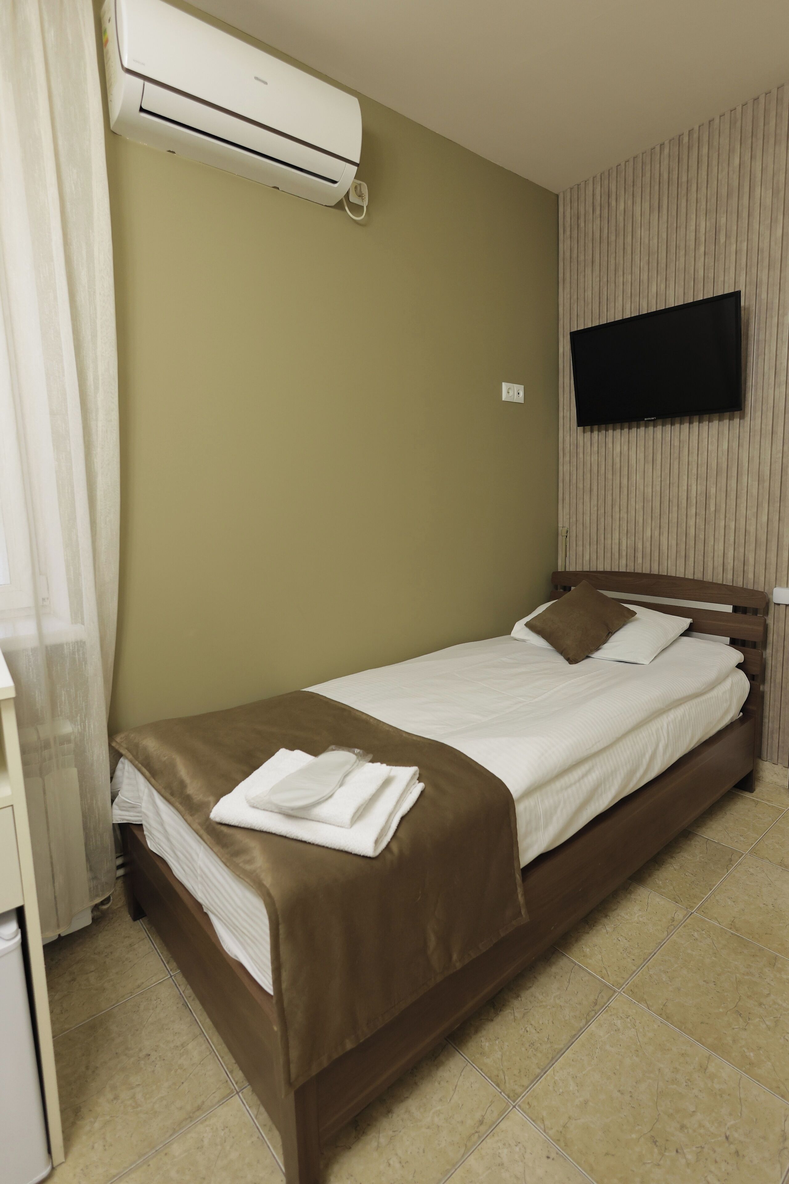 Una cama individual con cabecera marrón, un televisor de pantalla plana montado en la pared y un equipo de aire acondicionado sobre la cama.