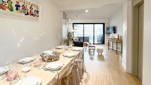 Un comedor moderno con una larga mesa preparada para una comida, un sofá y un televisor al fondo.