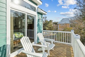 A wooden deck with white Adirondack chairs, a glass door leading to a covered area, and a white railing. - Dignified beachside home with private pool, golf cart, easy beach access (Emerald Isle)