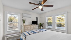 Quarto com cama, ventilador de teto, televisão, penteadeira e duas janelas com persianas.