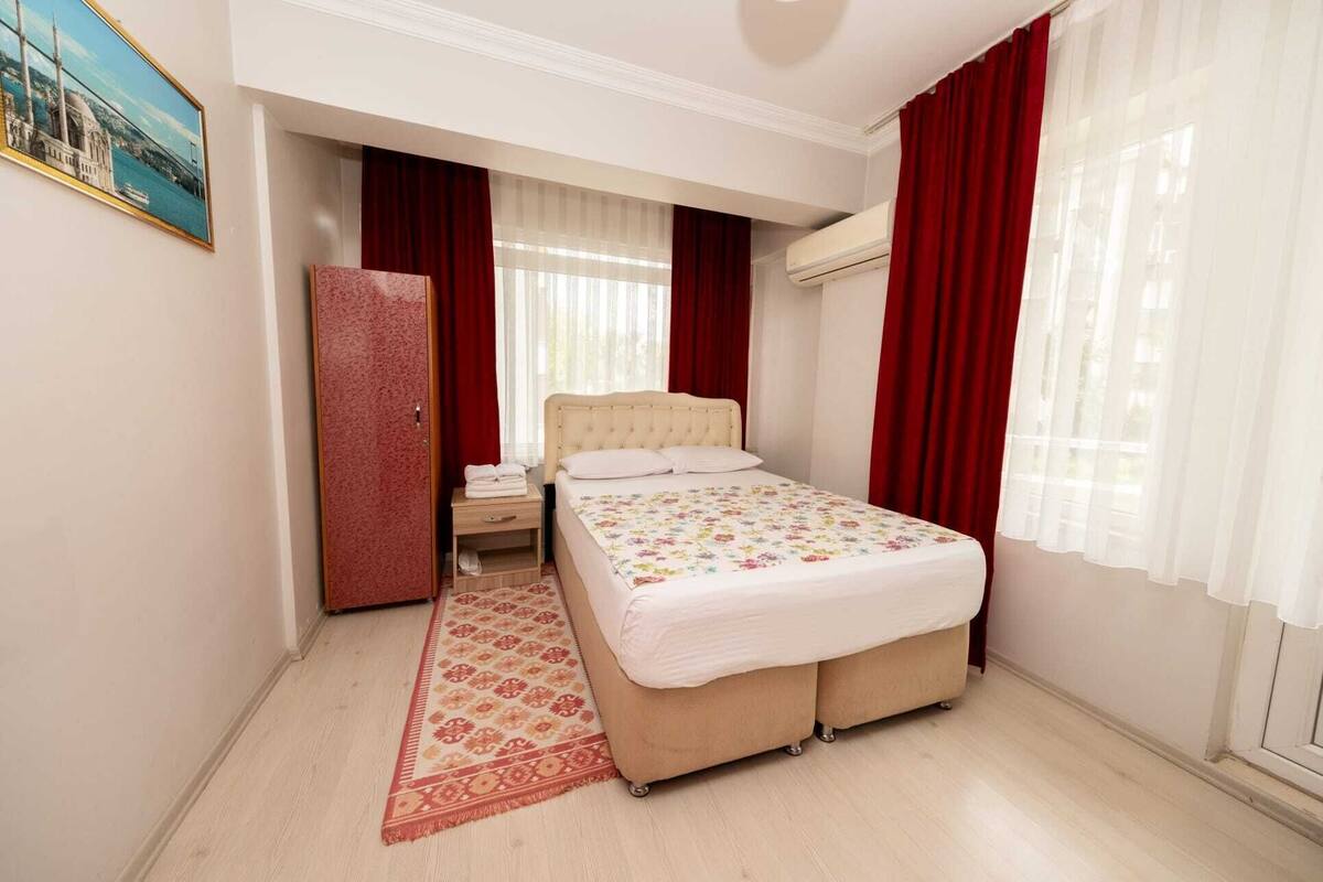 A hotel room with a bed, a nightstand, a red curtain, a window with sheer curtains, and a red carpet.