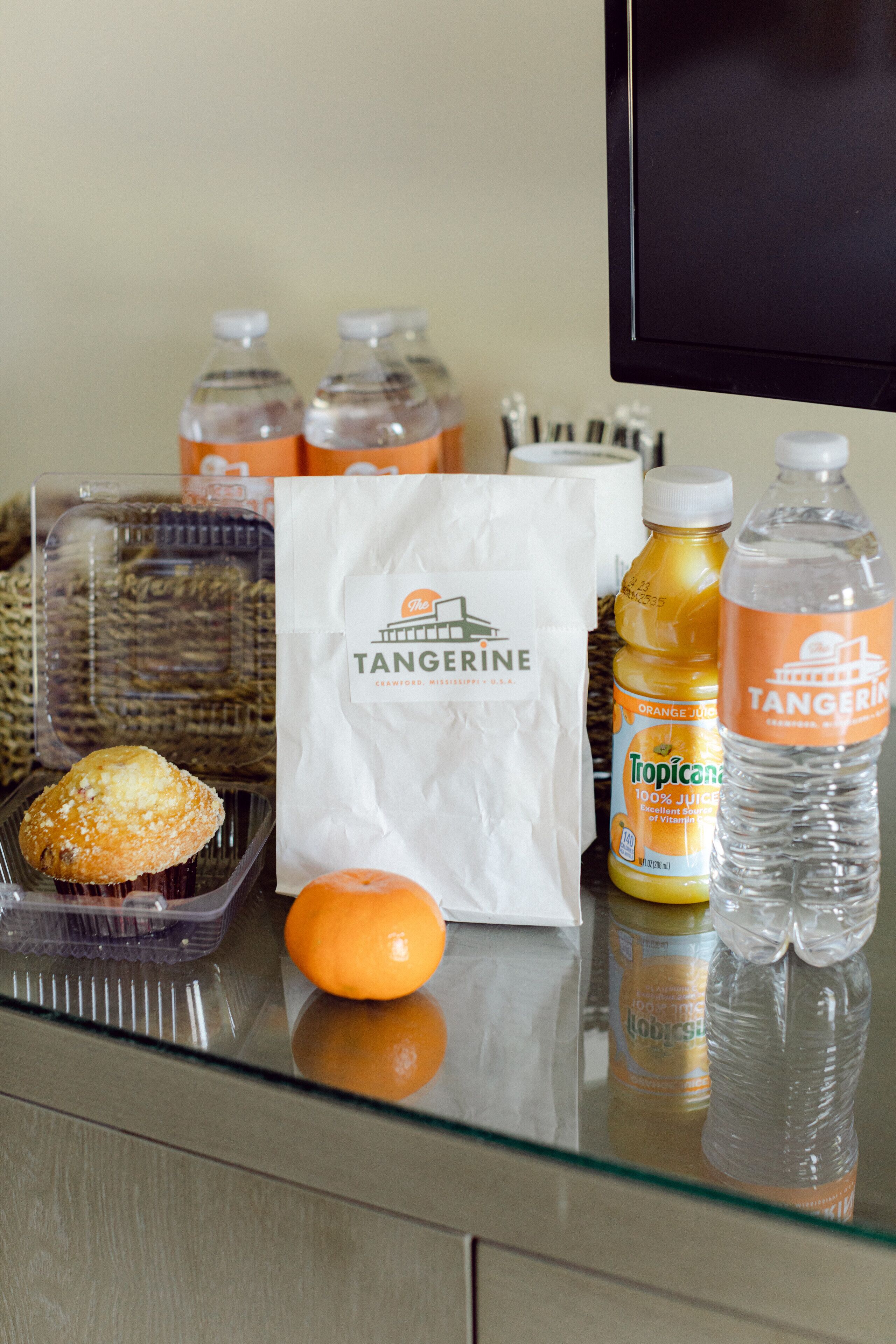 A glass table with a muffin, an orange, a bag labeled 'Tangerine', and two bottles of orange juice.