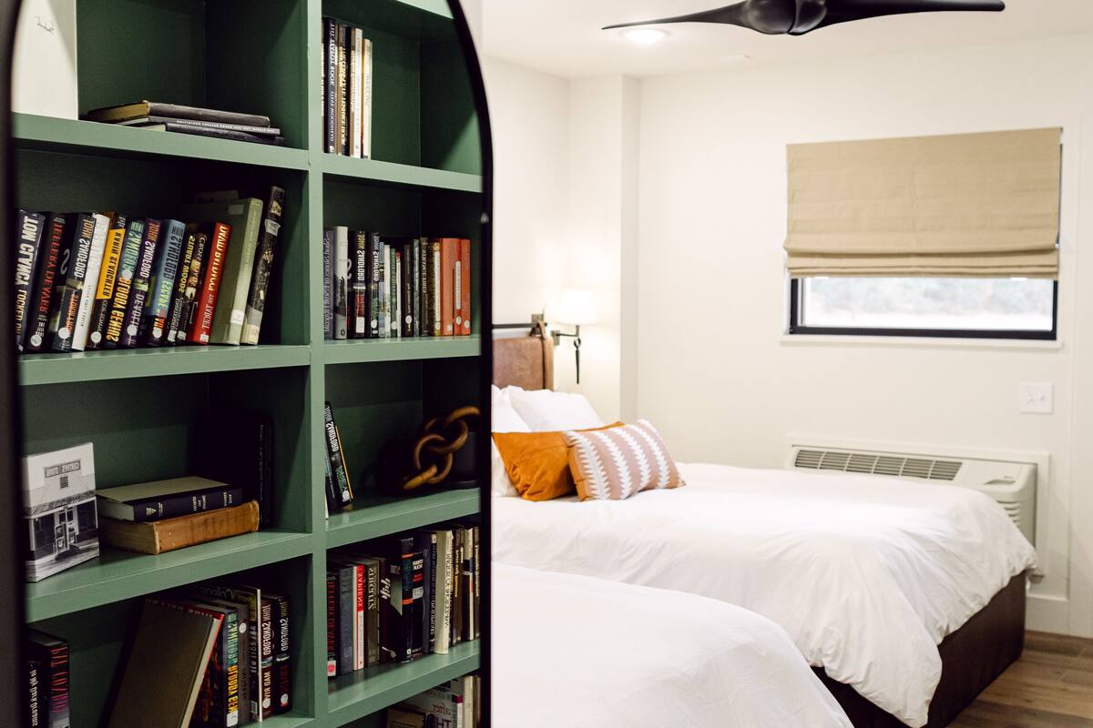 A bedroom with two beds, a green bookshelf, and a ceiling fan.