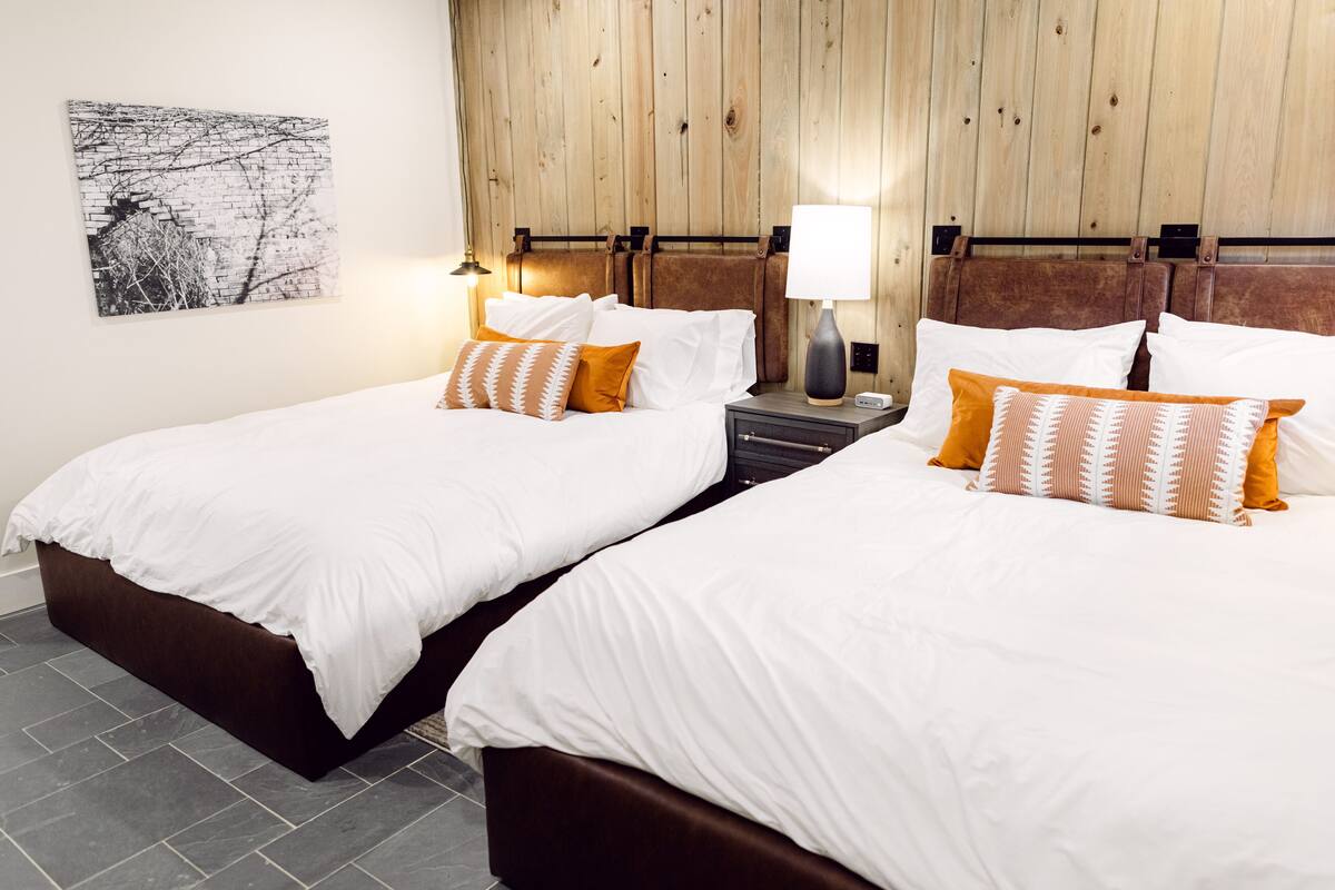 A hotel room with two beds, wooden headboards, a nightstand with a lamp, and a wall-mounted artwork.