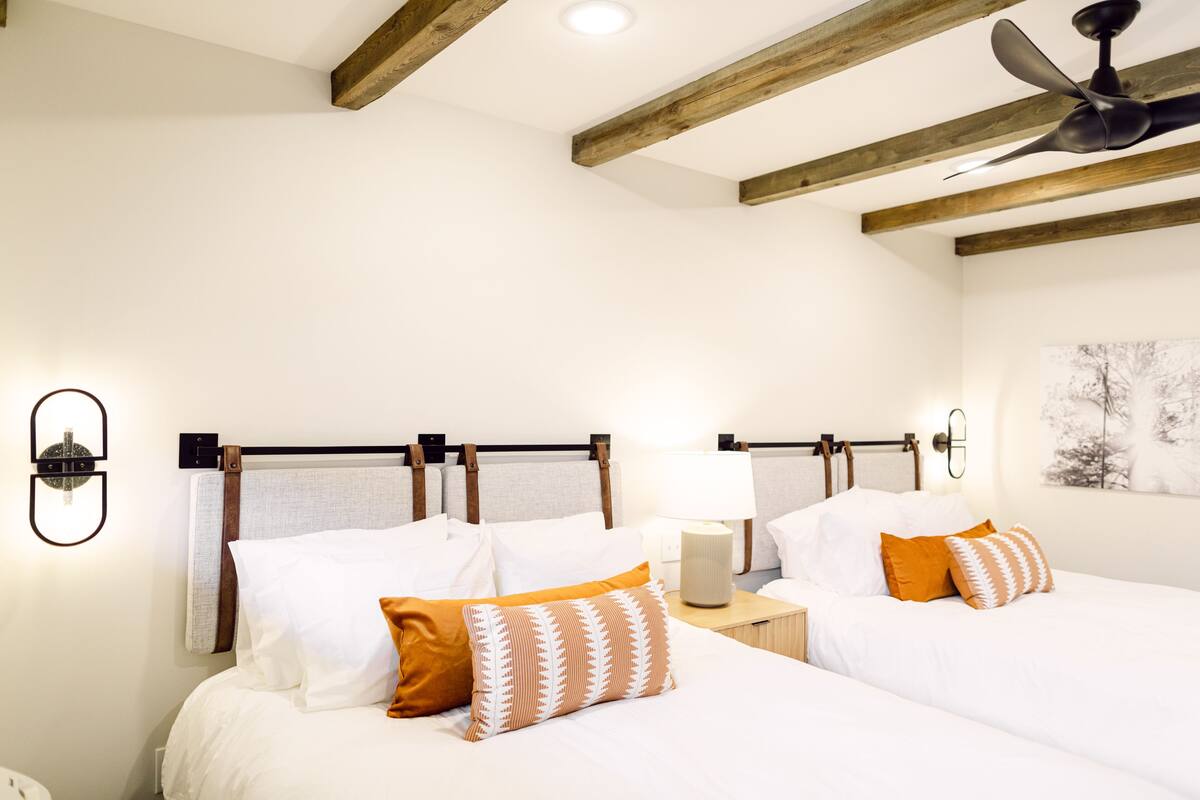 A hotel room with two beds, wooden beams on the ceiling, a ceiling fan, and a wall-mounted lamp.