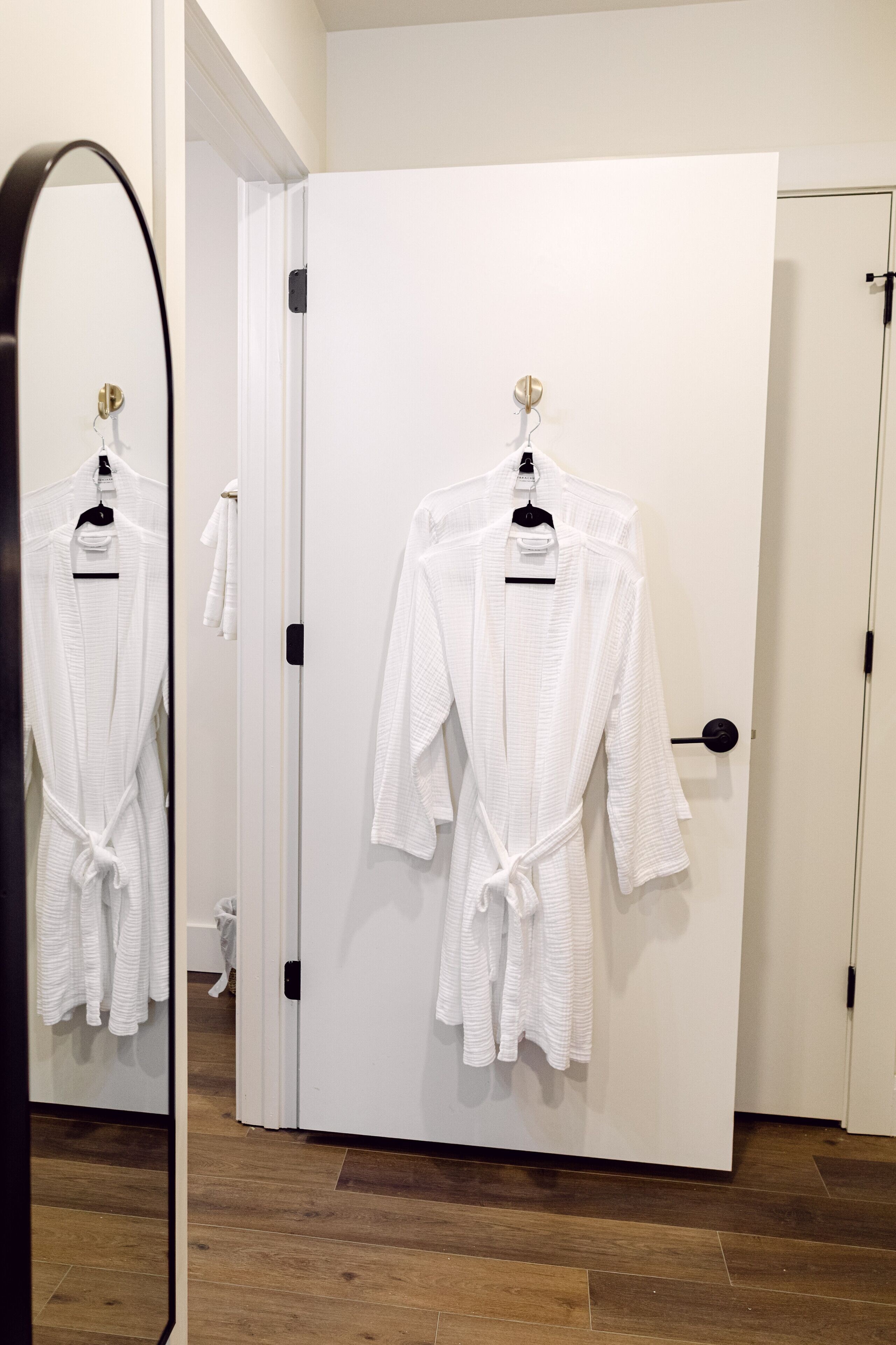Una habitación de hotel con piso de madera, una puerta blanca y dos batas blancas colgadas en ganchos.