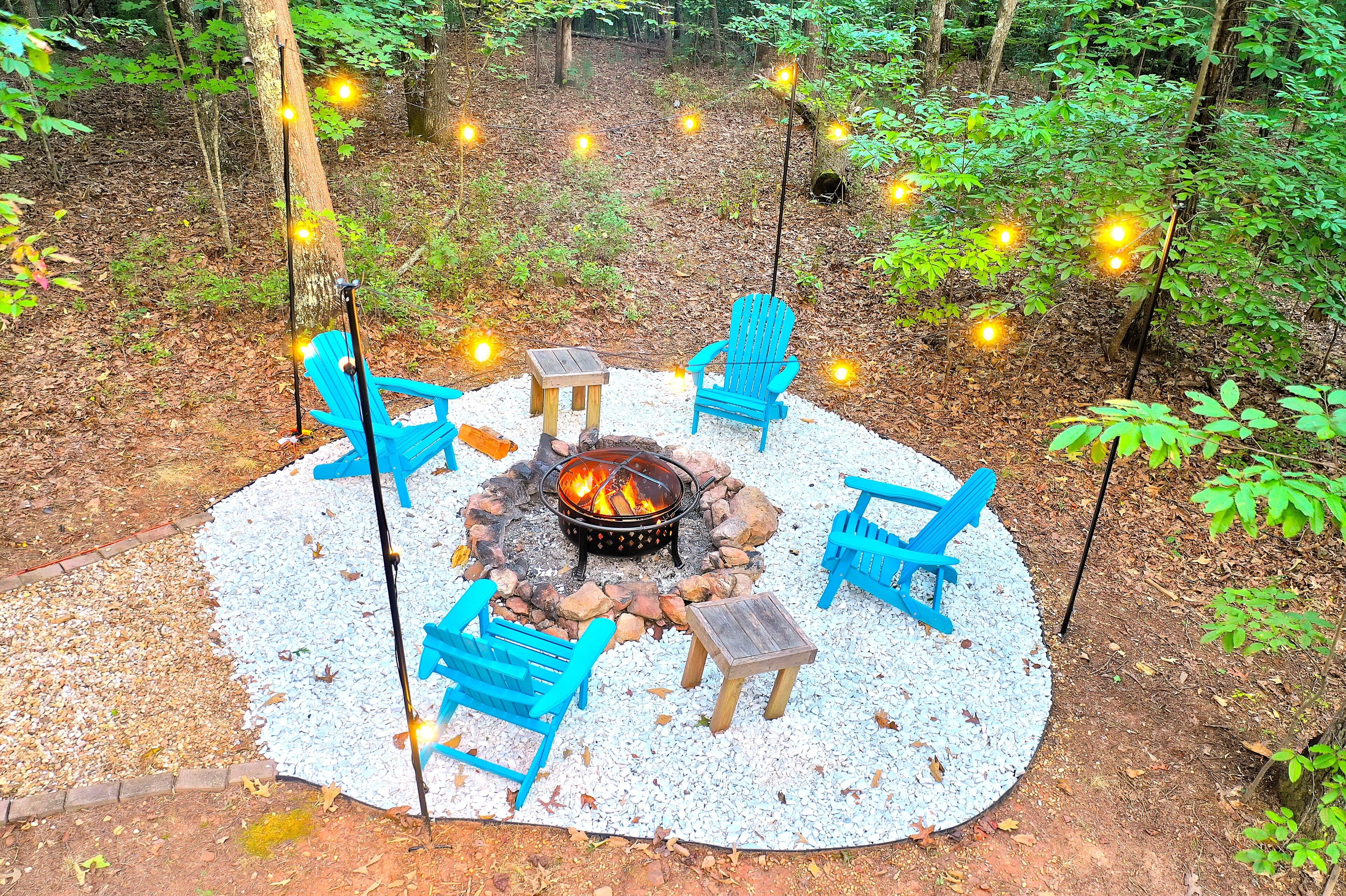 A circular outdoor seating area with a fire pit, surrounded by blue Adirondack chairs and a wooden table, set on a gravel surface in a forested area.