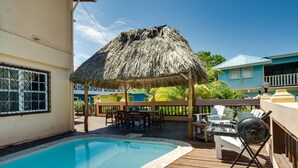 Una piscina con muelle de madera, un quiosco techado de paja, mobiliario exterior y una parrilla.