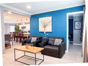Una sala de estar con pared azul, una mesa de centro de madera, dos sofás de cuero con cojines estampados, una zona de comedor con mesa y sillas de madera, y una cocina al fondo.