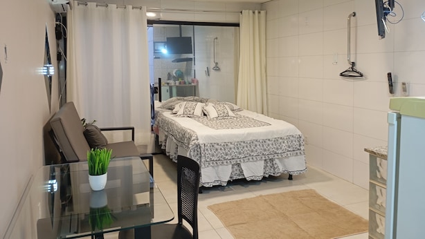Een knus slaapkamer met een bed, een klein tafeltje met een plant, een stoel, en een douchegedeelte zichtbaar door een glazen deur.