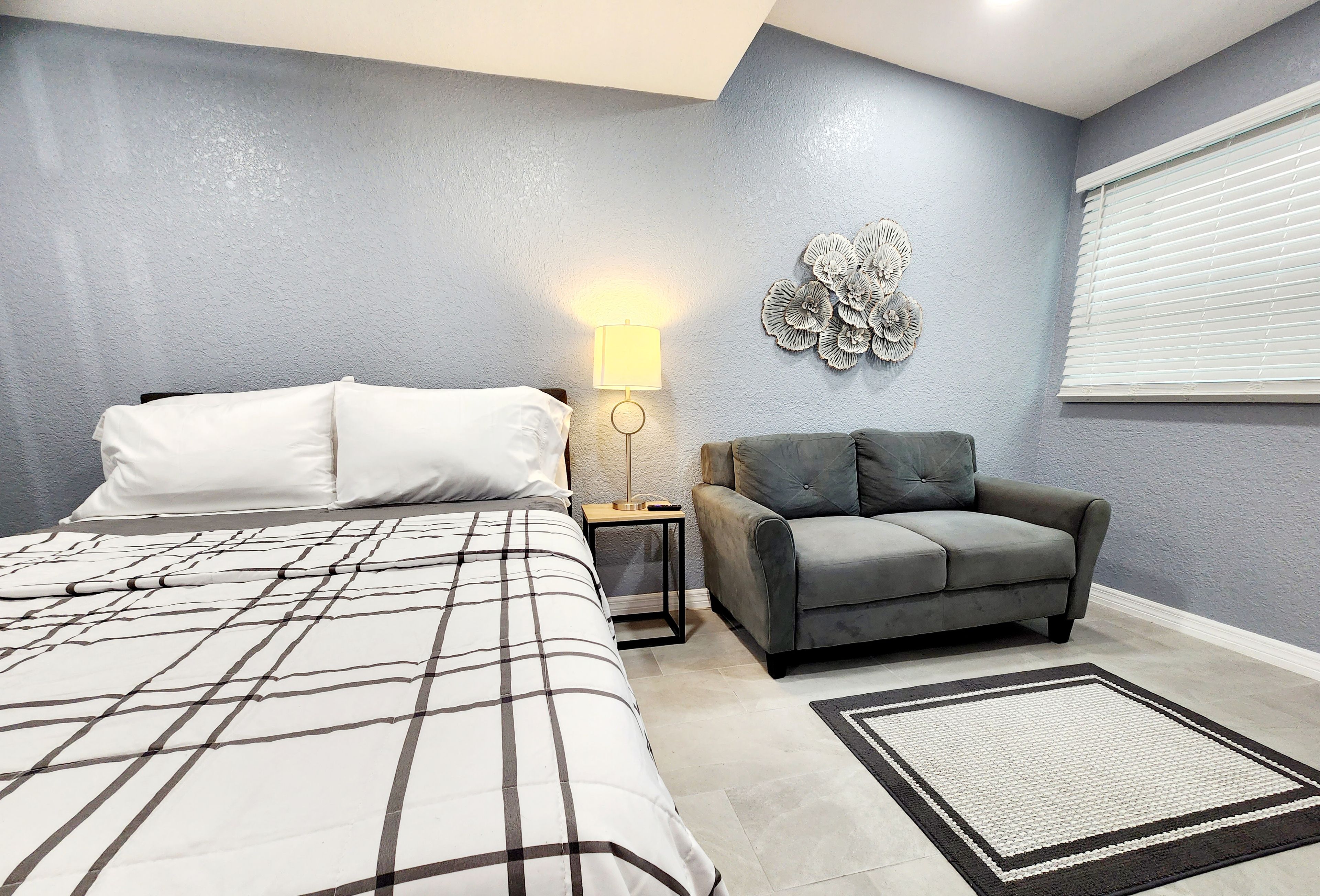Un dormitorio con una cama, un sofá gris, una mesita de noche con lámpara y un adorno en la pared.