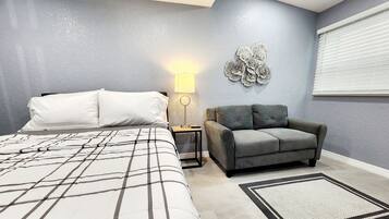 Un dormitorio con una cama, un sofá gris, una mesita de noche con lámpara y un adorno en la pared.