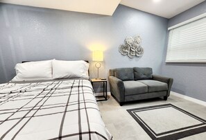 Un dormitori amb un llit, un sofà gris, una tauleta de nit amb una làmpada i una decoració mural.