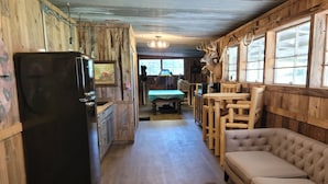 A rustic cabin interior with a wooden dining table, a sofa, a refrigerator, and a mounted deer head.