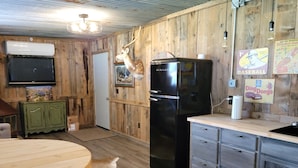 A rustic kitchen with wooden walls, a black refrigerator, a wooden dining table, and a television mounted above the cabinet.