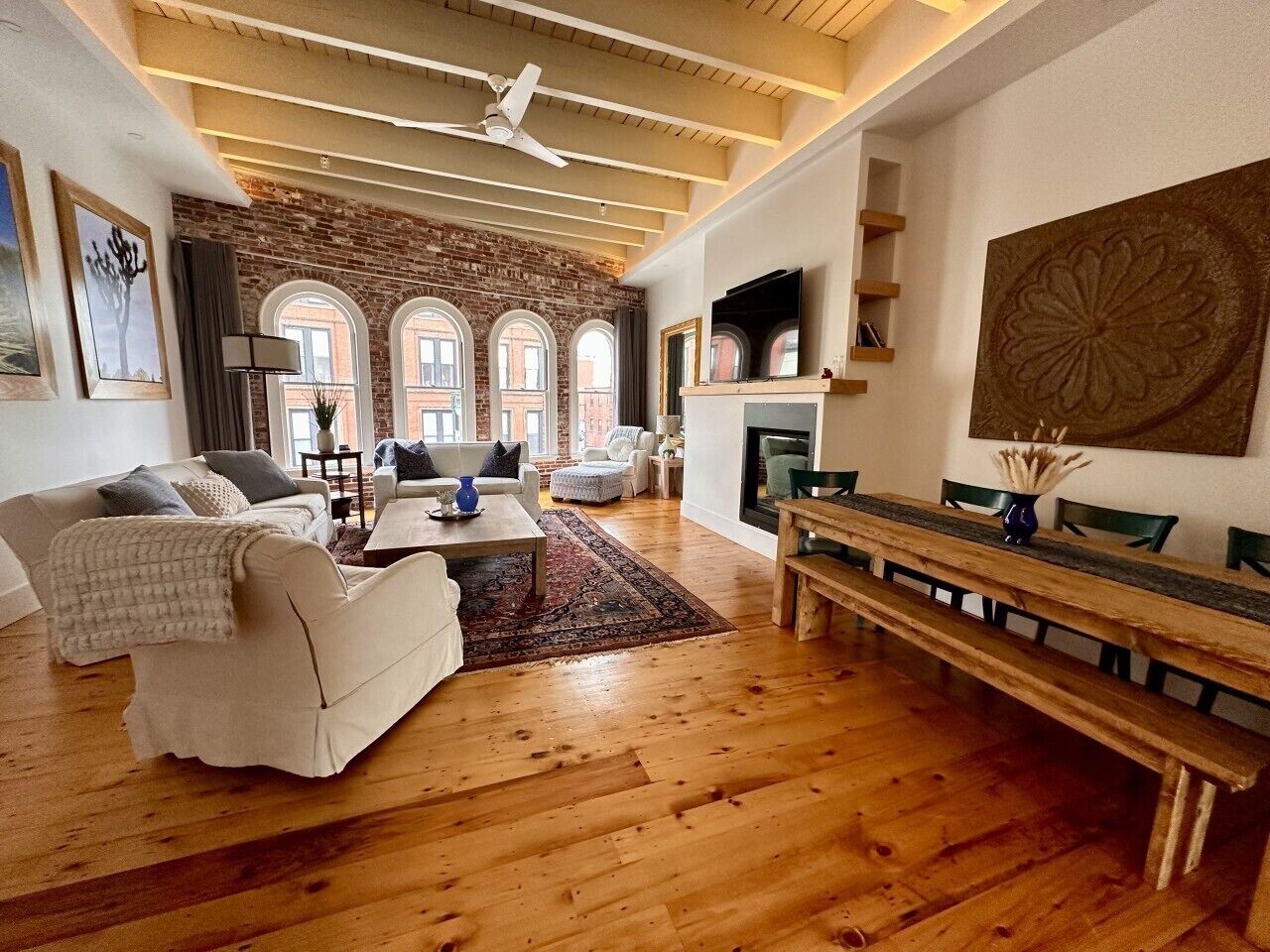 A spacious living room with a fireplace, wooden flooring, and a large window overlooking a brick wall.