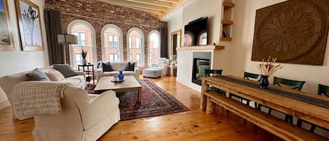 A spacious living room with a fireplace, wooden flooring, and a large window overlooking a brick wall.