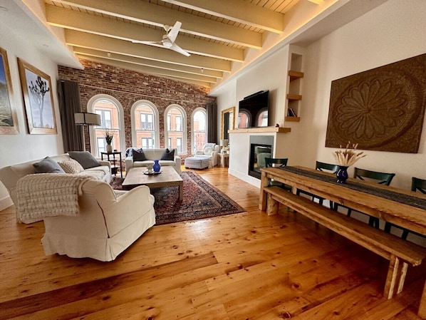 A spacious living room with a fireplace, wooden flooring, and a large window overlooking a brick wall.