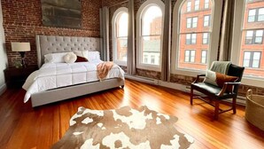 A bedroom with a large bed, a leather chair, and a view of buildings through arched windows. - Stunning Luxury Apt/Old Port Arts Dist/Sleeps8 (Portland)