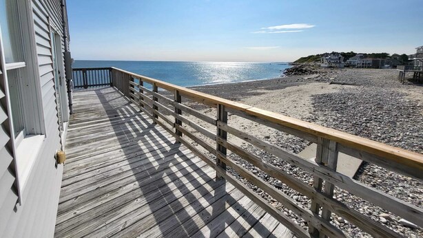 Una terraza de madera con barandilla que da a una playa de guijarros y al océano.
