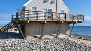 Una casa con una terraza de madera que se extiende sobre una playa rocosa, una escalera que sube a la terraza, y vistas al océano.