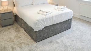 Una cama bien hecha con sábanas y toallas blancas, un cabecero gris y lámparas de noche sobre una mesita de noche gris.