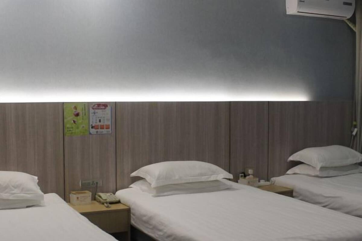 A hotel room with two single beds, a wooden headboard, a bedside table with a lamp, and a wall-mounted air conditioner.